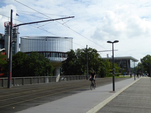 Drewniane torowisko i rowerzyści na tle budynku Europejskiego Trybunału Praw Czowieka.