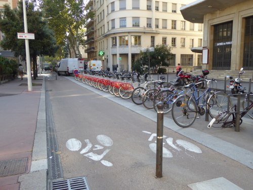 Stacja rowerów publicznych i stojaki przy wydzielonej drodze rowerowej.