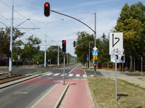 Znak informujący o sposobie skręcenia rowerem w lewo. Na zdjęciu jest widoczny mały trójkomorowy sygnalizator rowerowy.
